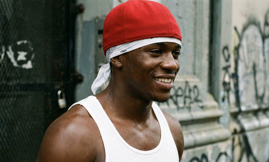 Smiling young man on a city street, wearing a white bandana under a red baseball cap and a casual tank top.